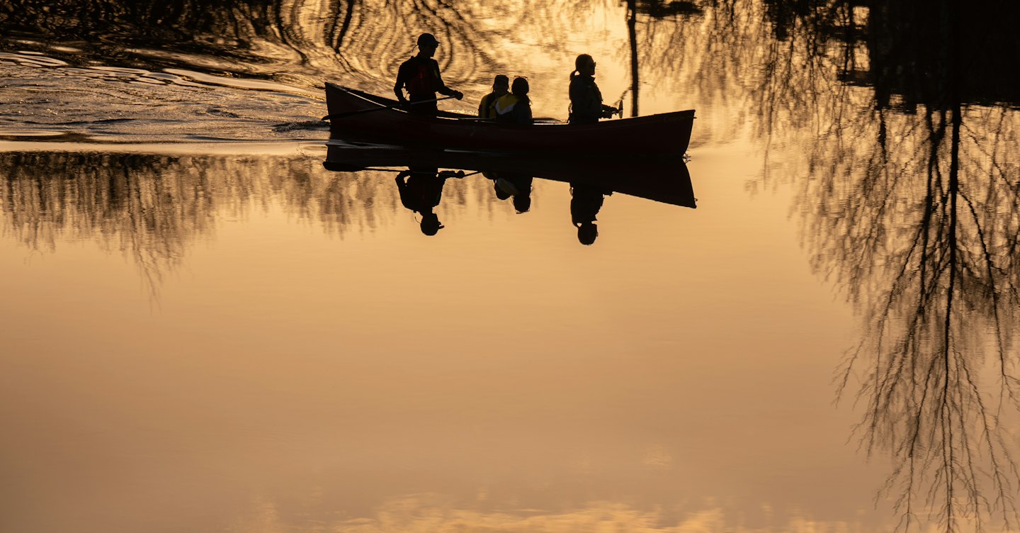 Three people paddle a red canoe silhouetted against a sunset.